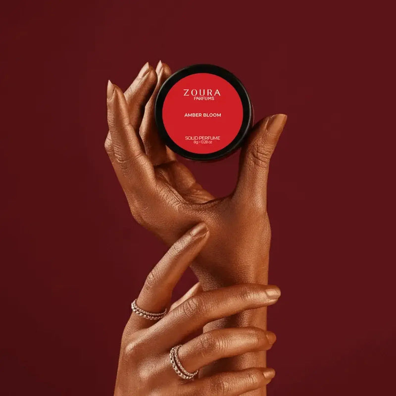 Hands holding Zoura Amber Bloom Unisex Solid Perfume jar against red background