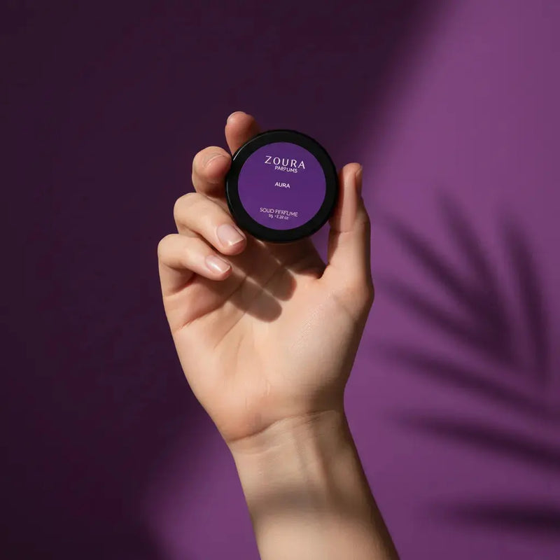 Hand holding Aura Solid Unisex Perfume tin by Zoura Parfums against purple backdrop